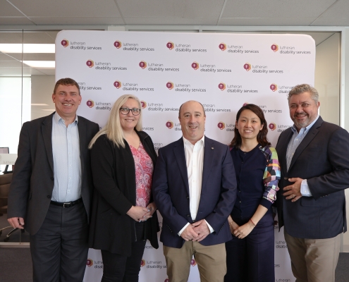 Group photo of the five executive staff at Lutheran Disability Services, standing in the office infront of a LDS branded backdrop with the logo tiled repeatedly.