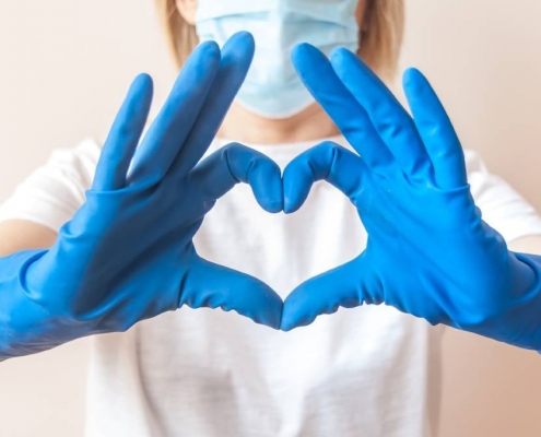 image of woman with face mask, wearing a white shirt, making a heart shape with her hands wearing blue rubber gloves.