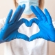 image of woman with face mask, wearing a white shirt, making a heart shape with her hands wearing blue rubber gloves.