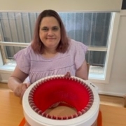 Client Samantha in a purple shirt and purple hair smiles at the camera whilst using her beanie maker to make a read beanie. She is sitting at a wooden table in the living area.